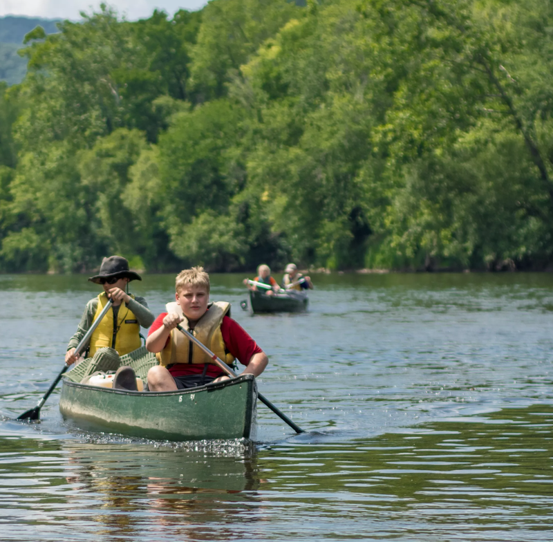 The image shows two young people paddling a green canoe on a river. The person in the front is wearing a red life jacket and holding an oar, while the person in the back is wearing a yellow life jacket and a hat. There are other canoes with people in the background, and the river is surrounded by green trees. The water is calm and reflects the sky and trees.